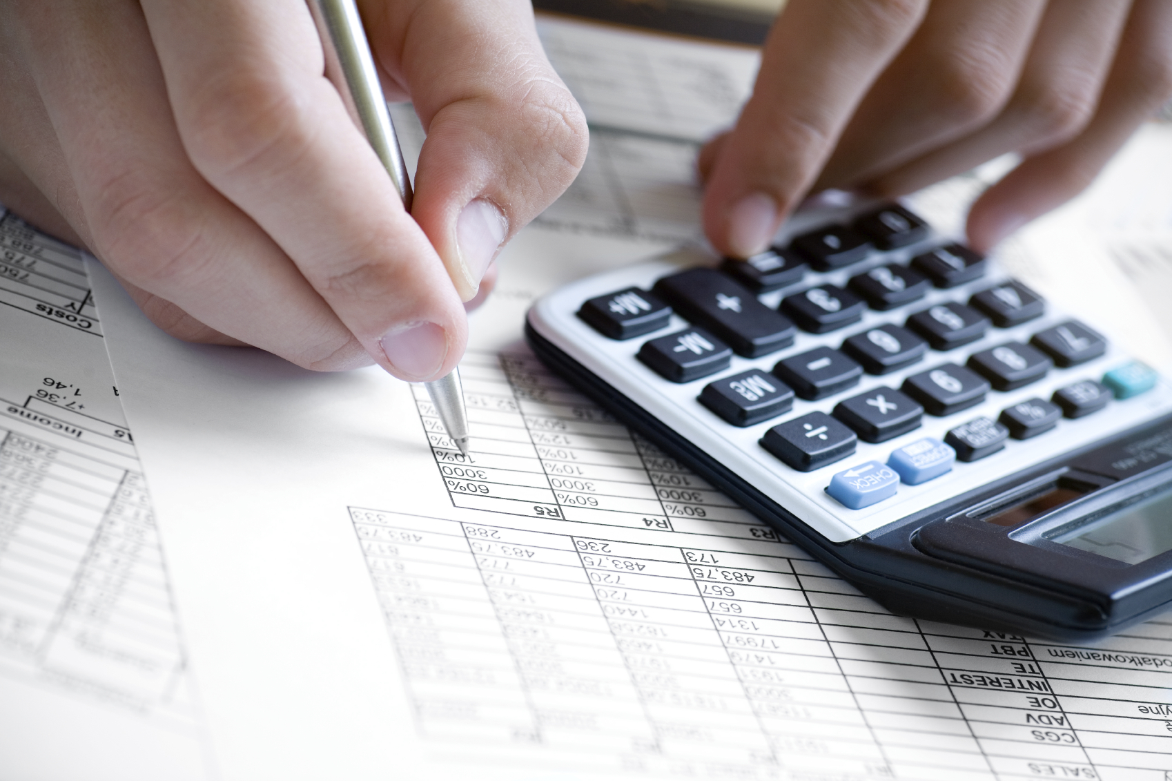 Hand holding a pen checking a balance sheet on a calculator.