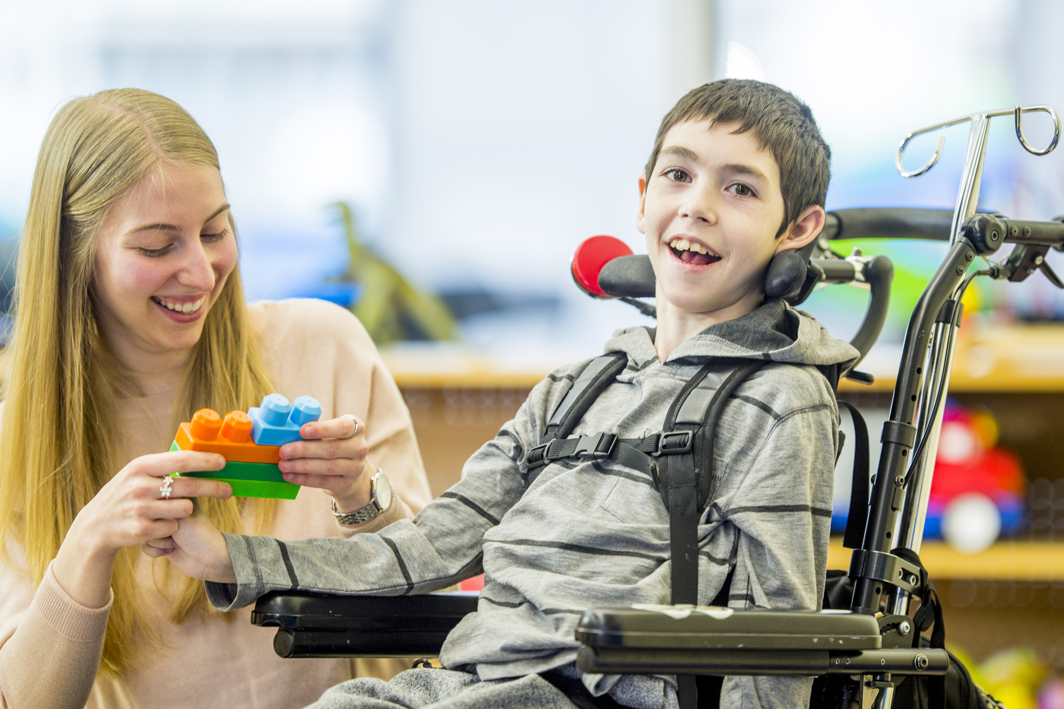 Young boy in wheelchair smiling. He is with a young female carer who is holding colourful building blocks.