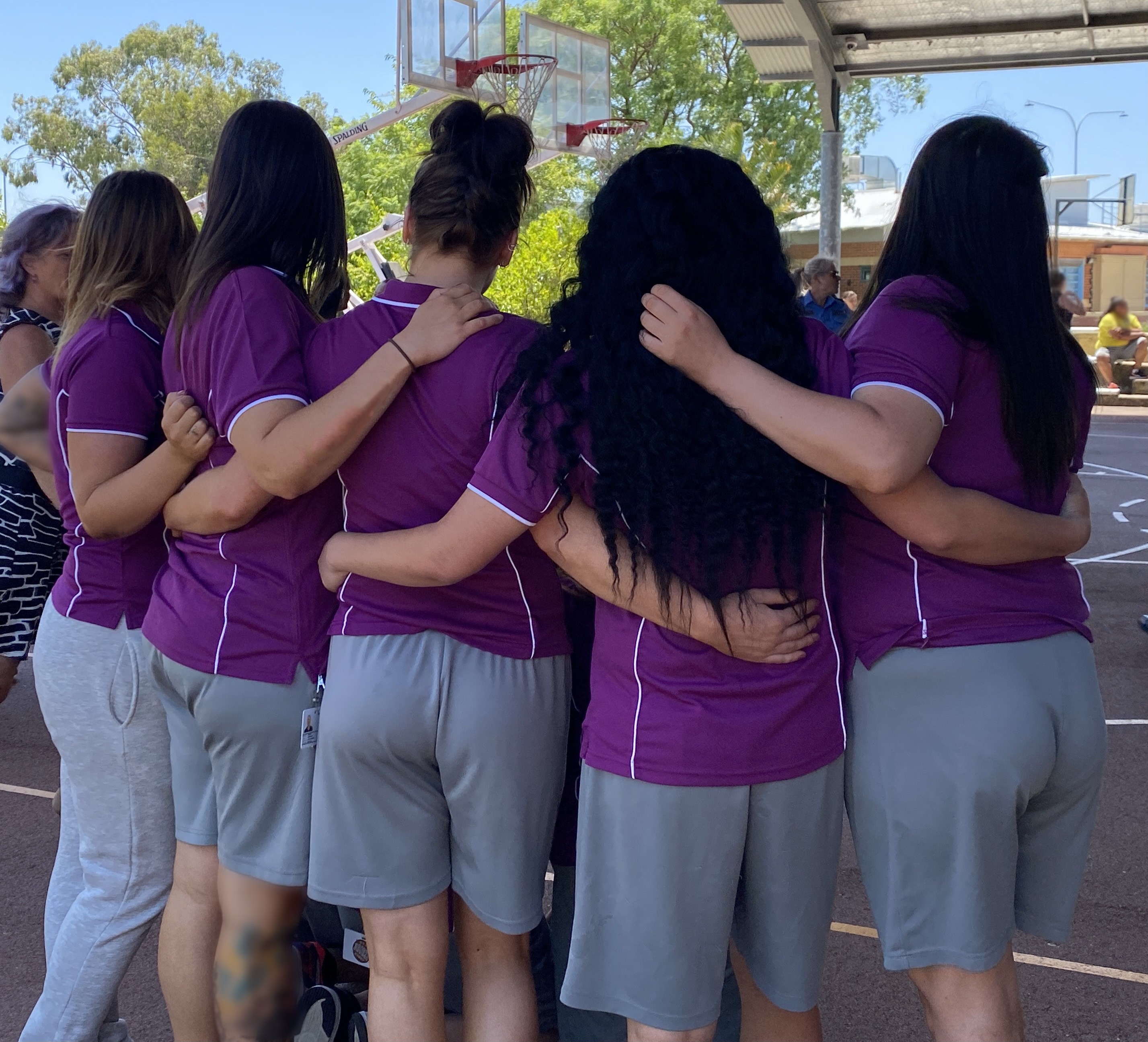 A group of women wearing purple tops and grey shorts are standing in a group with their arms around each other. The women have dark hair and are standing outside on a sunny day with blue sky.