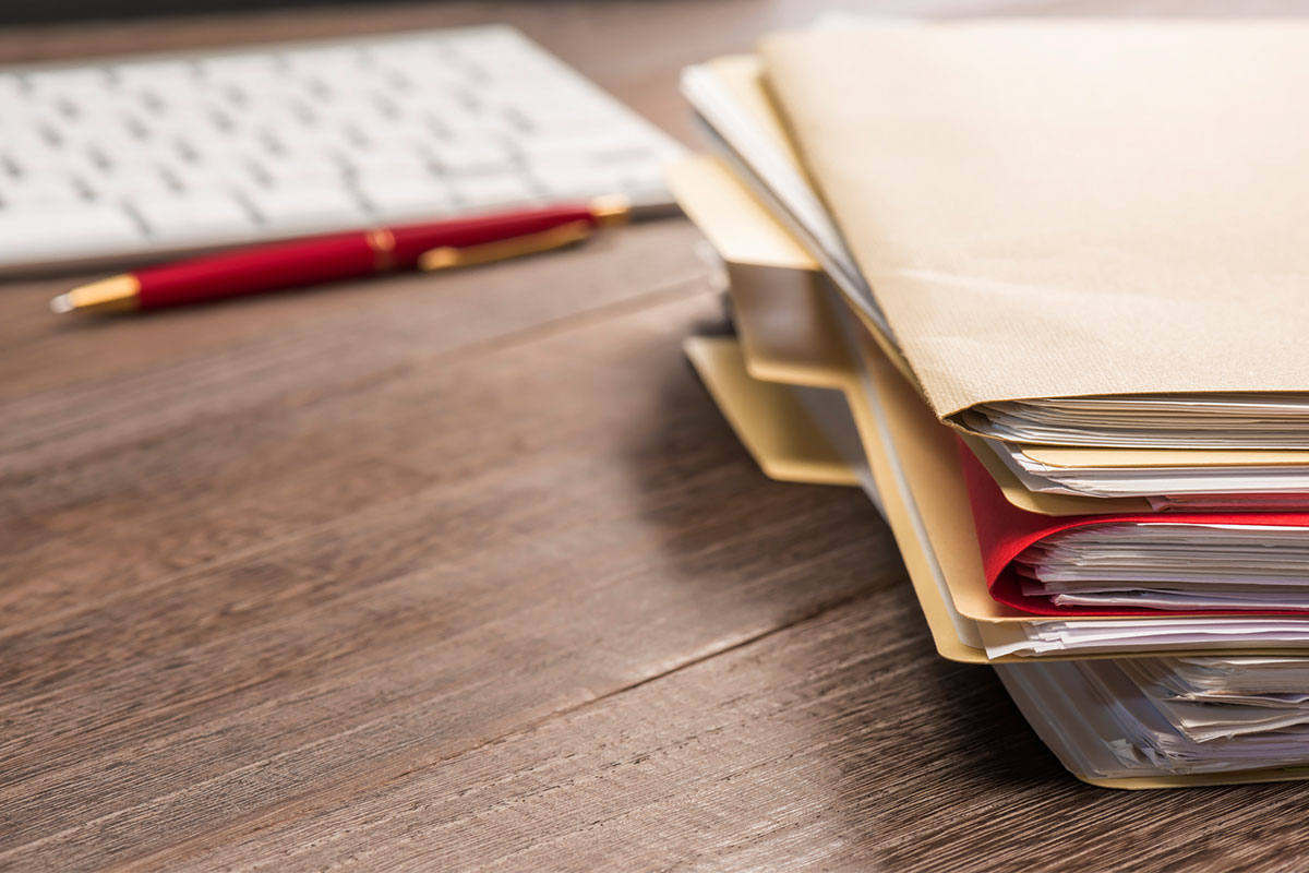 A stack of paper files, and pen and a computer keyboard on a desk.