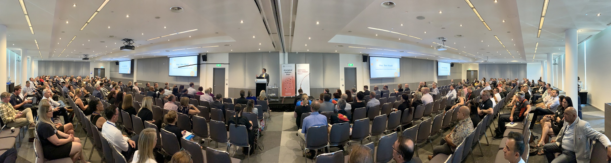 A panoramic photo of a seated crowd listening to a speaker presenting information.