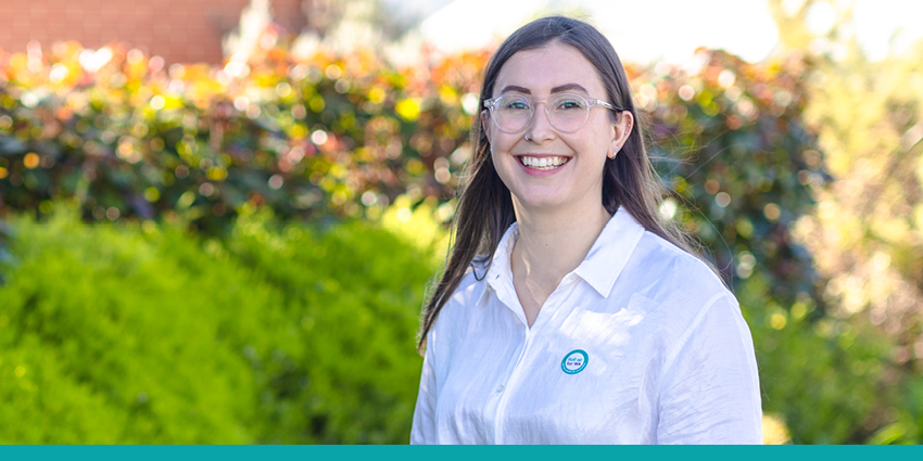 Brown haired girl in glasses and white top with a roll up for WA sticker on her shirt standing in front of green hedges.