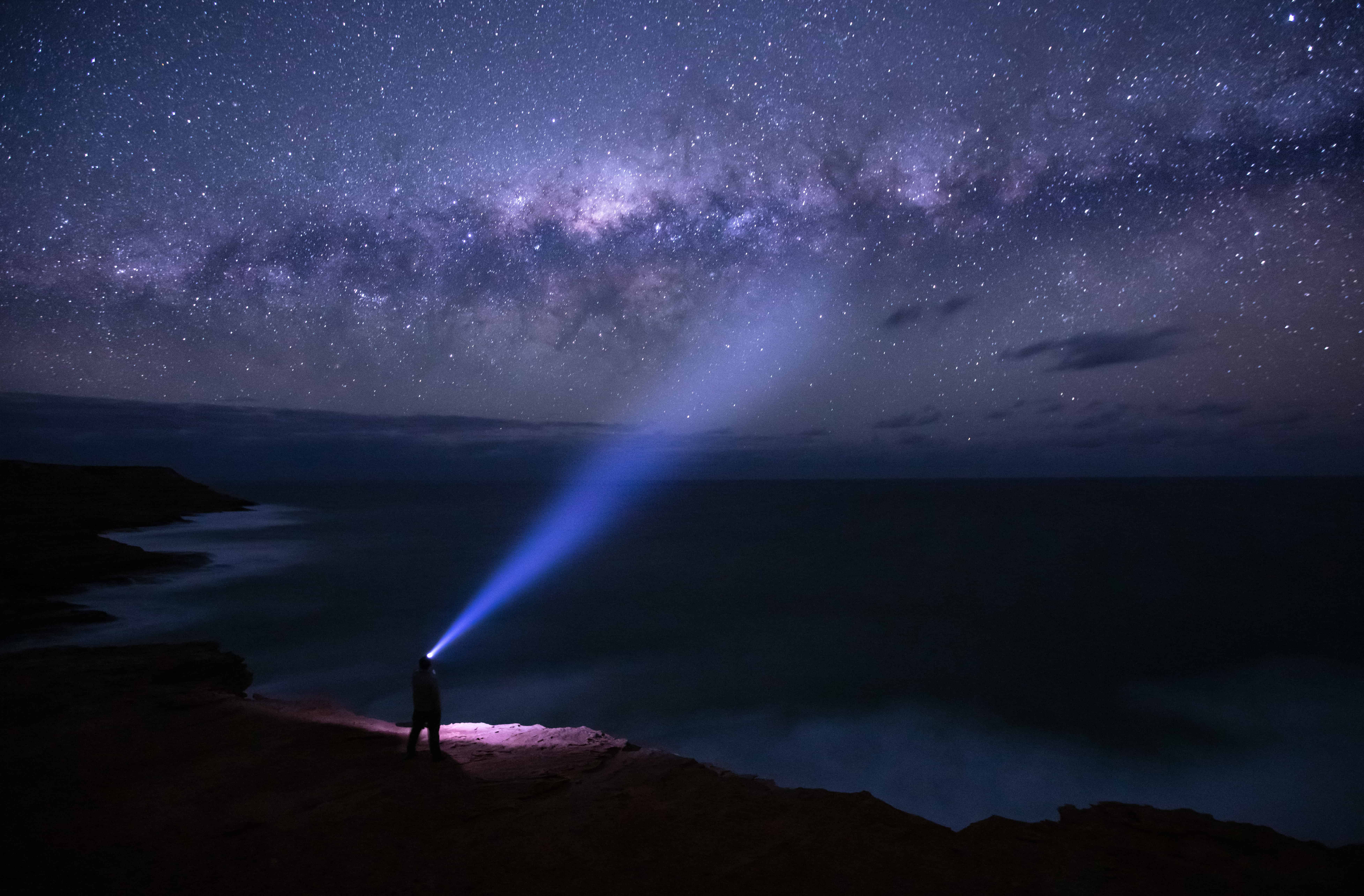 Person with a headlamp standing on the edge of a cliff looking out at the stars in the night sky