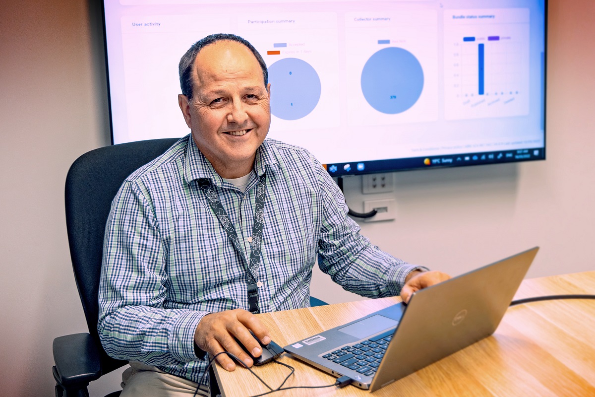 A man sitting on a laptop with at a desk with powerpoint presentation behind him