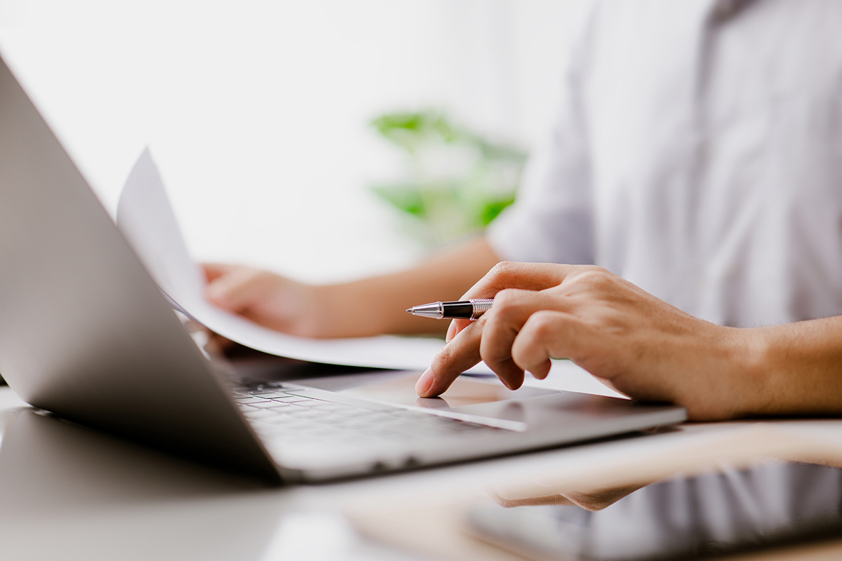 A close up of a hand holding a pen and paper, while navigating a laptop computer.