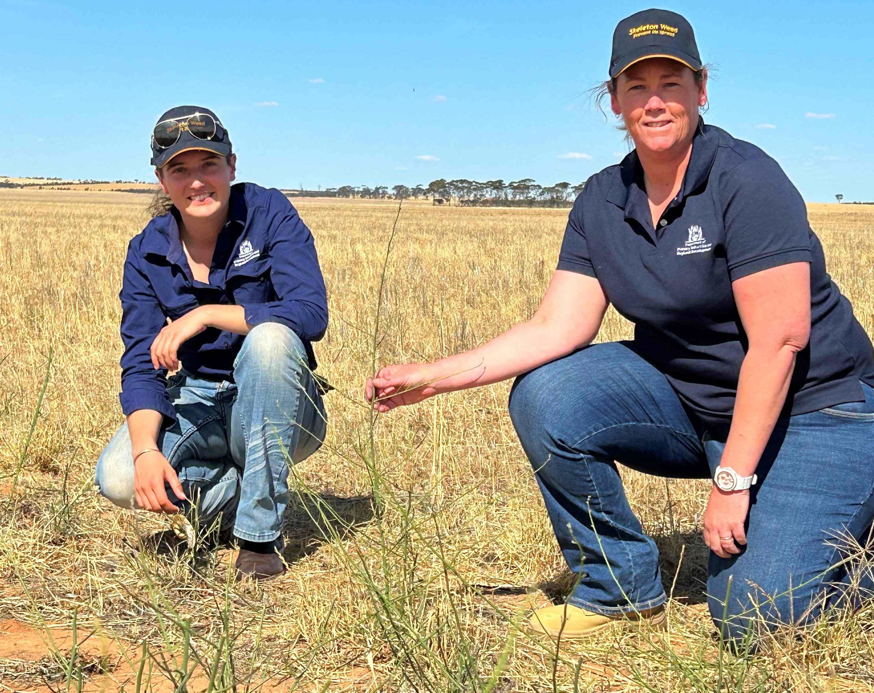 Two women in a dry paddock with a small green plant.