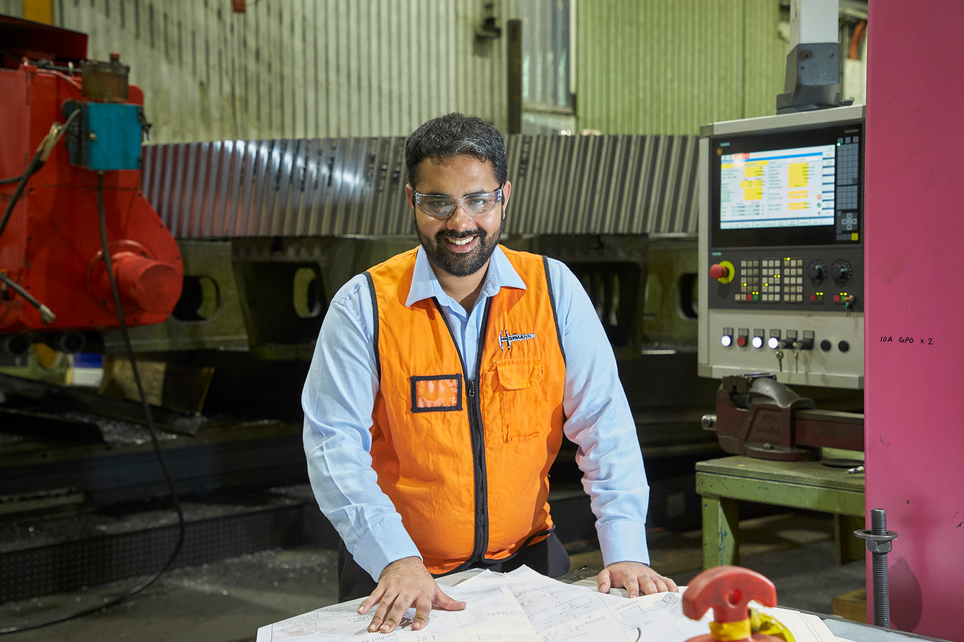 An engineer wearing an orange vest and protective eyewear, in a warehouse, smiling at the camera.