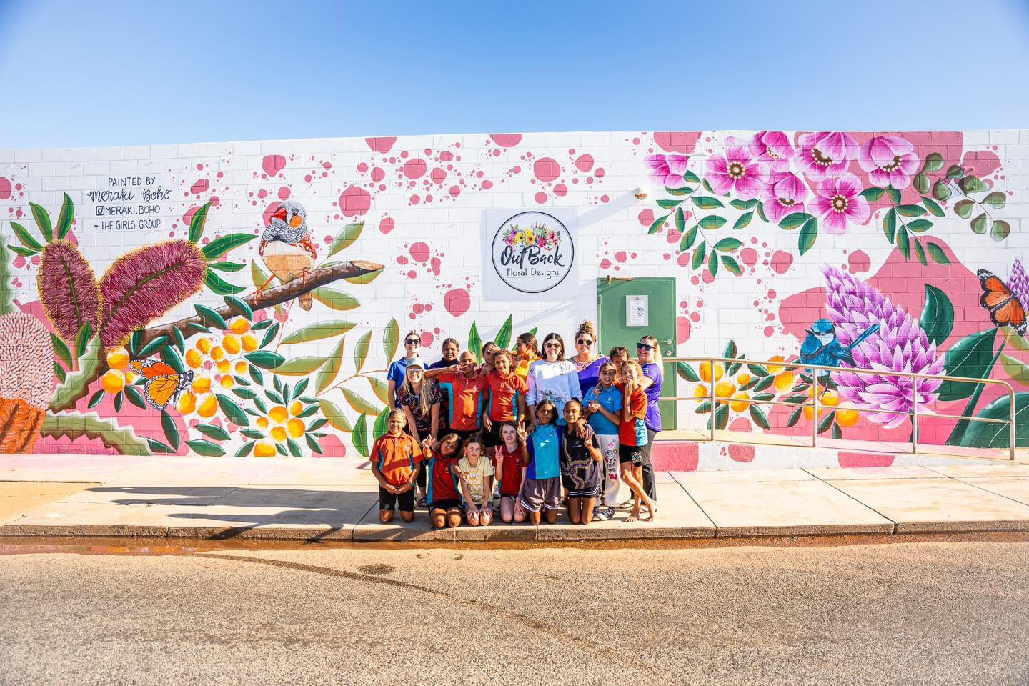 group photo of a group of Aboriginal girls and women on a street with a colourful murel on a wall behind them