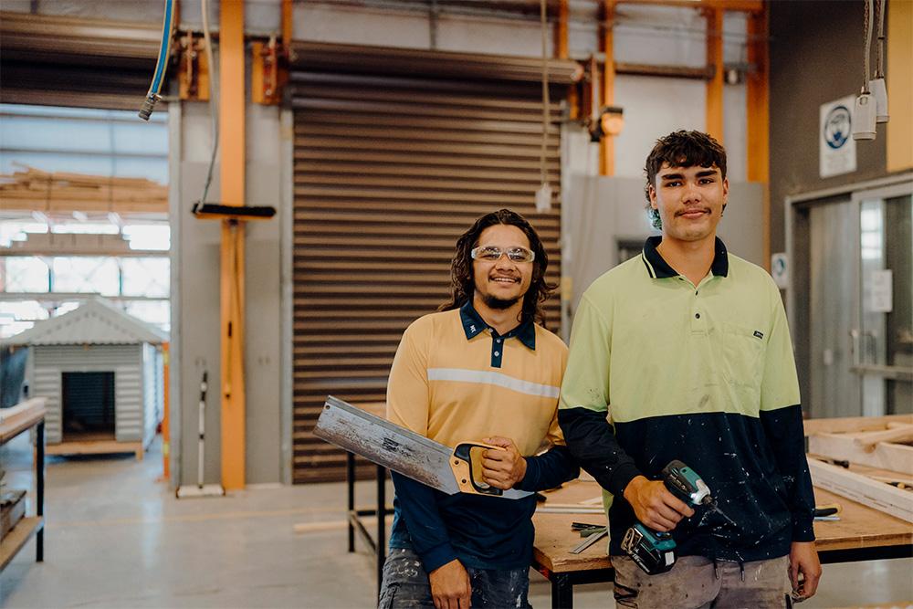 Two young male apprentices in work attire, smiling in a workshop.