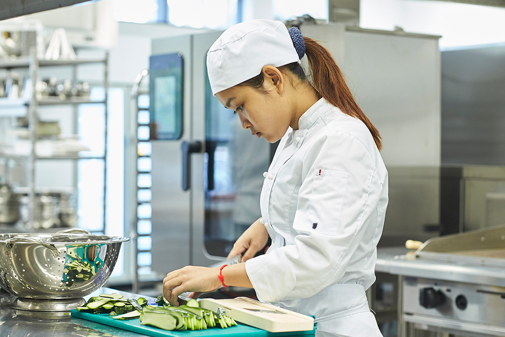 A young female student chef in a white uniform and hat preparing vegetables in a commercial kitchen. 