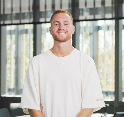 A man wearing a white t-shirt stands by a window smiling