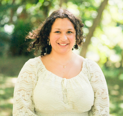 A woman in a white blouse stands with trees in the background