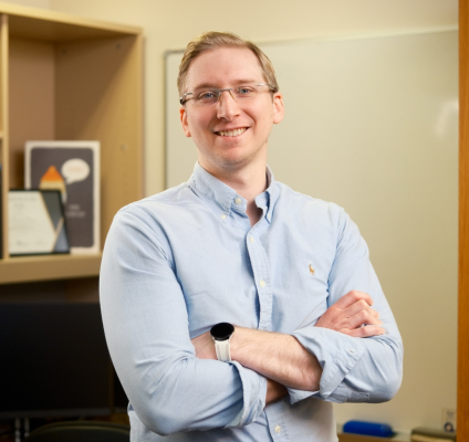 a man in a pale blue shirt bespectacled stands before a desk