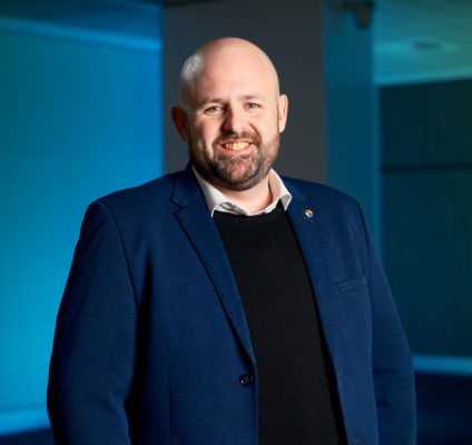  A bald man in a blue suit poses against a lush green wall, showcasing a professional appearance.