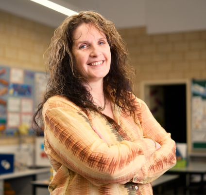 A woman with long hair stands in a classroom, surrounded by desks 