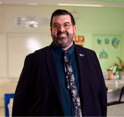 A man in a suit and tie stands confidently in a classroom, surrounded by desks and educational materials.