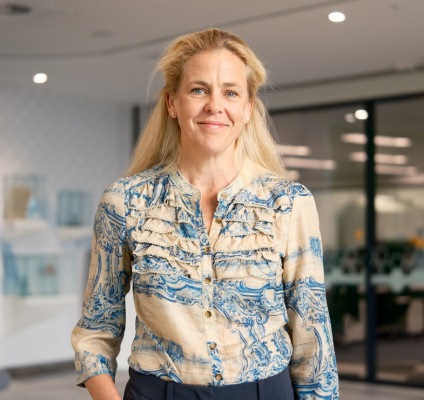 A woman wearing a blue shirt and navy pants stands confidently in a modern office environment.