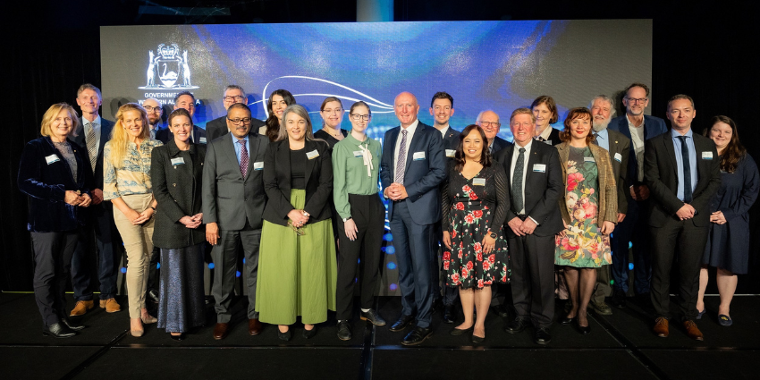 A diverse group of individuals stands together on a stage, smiling.