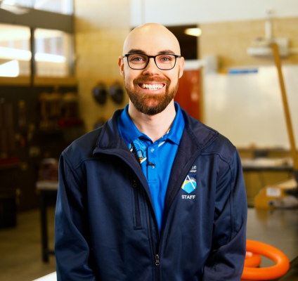 A man with glasses and a blue jacket poses in a classroom