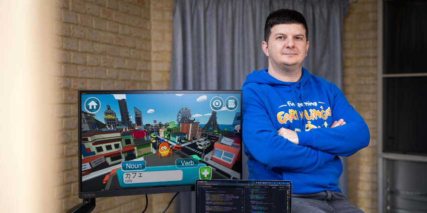 man in a blue hoodie with Earthlingo on it sitting next to a computer screen with a video game displayed and a laptop showing code