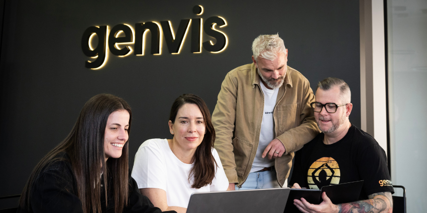 office workers in front of dark wall with genvis on it, discussing projects and looking at laptop