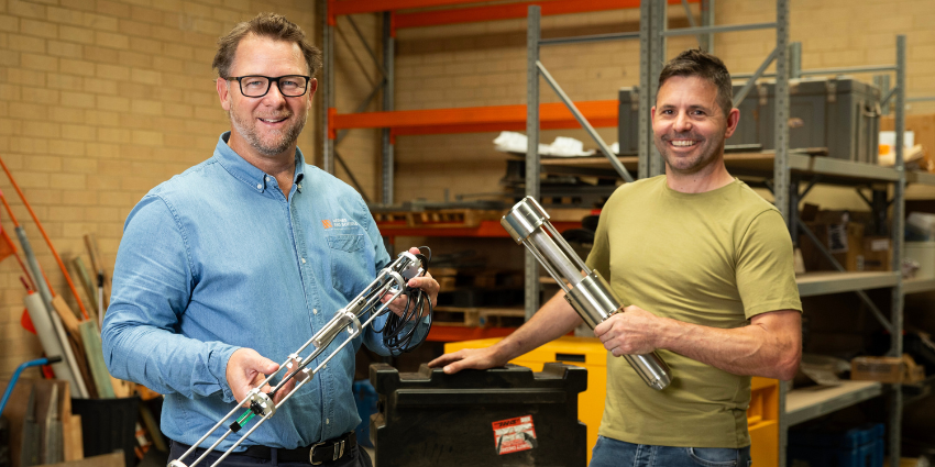 two men standing in industrial workspace holding machine parts and smiling