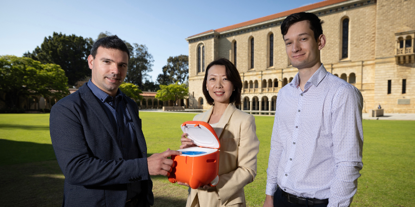 lady and two men standing on university campus lawn holding orange cube