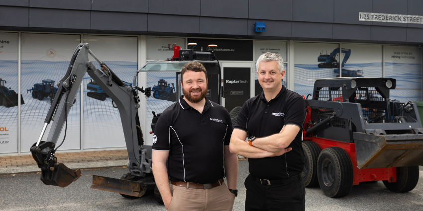 dark haired and grey haired man standing in front of fork lift and tractor