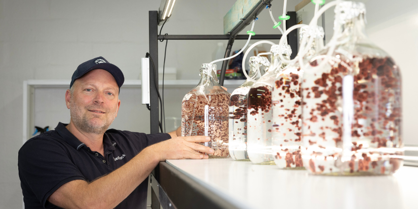 man in black polo with black cap on stands next to jars of liquid with red particles inside