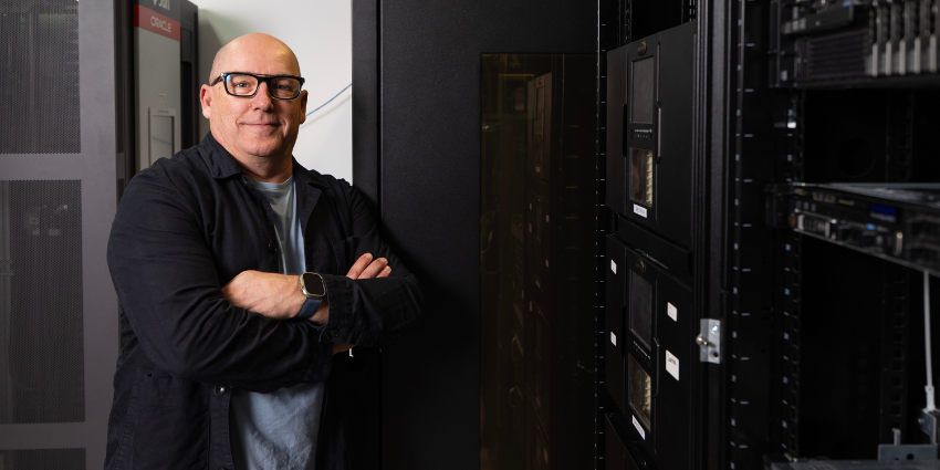 dark image, bald man with thick-rimmed black glasses standing next to black storage for tape records