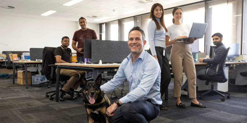 man in blue collared shirt smiling and kneeling next to dog with office workers at their desks in background