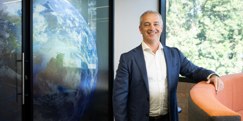 man in blue suit jacket stands in office environment with a projection of the earth behind him