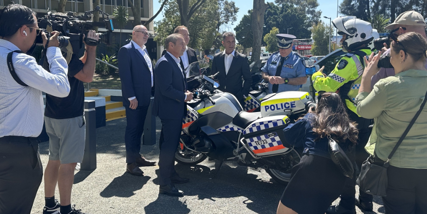 Public officials and the media grouped around a parked police motorcycle