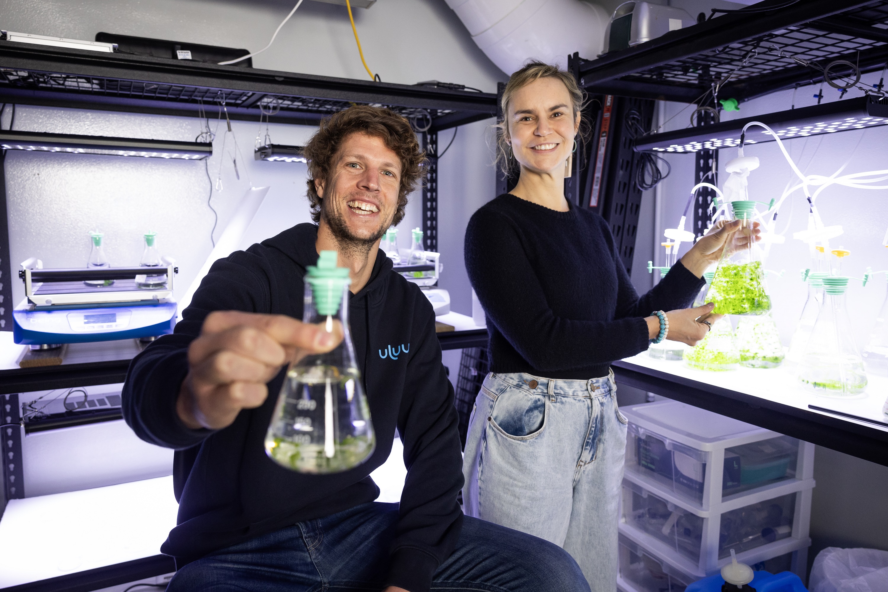 two young scientists, one male, one female in lab holding test tubes and vials with green liquid