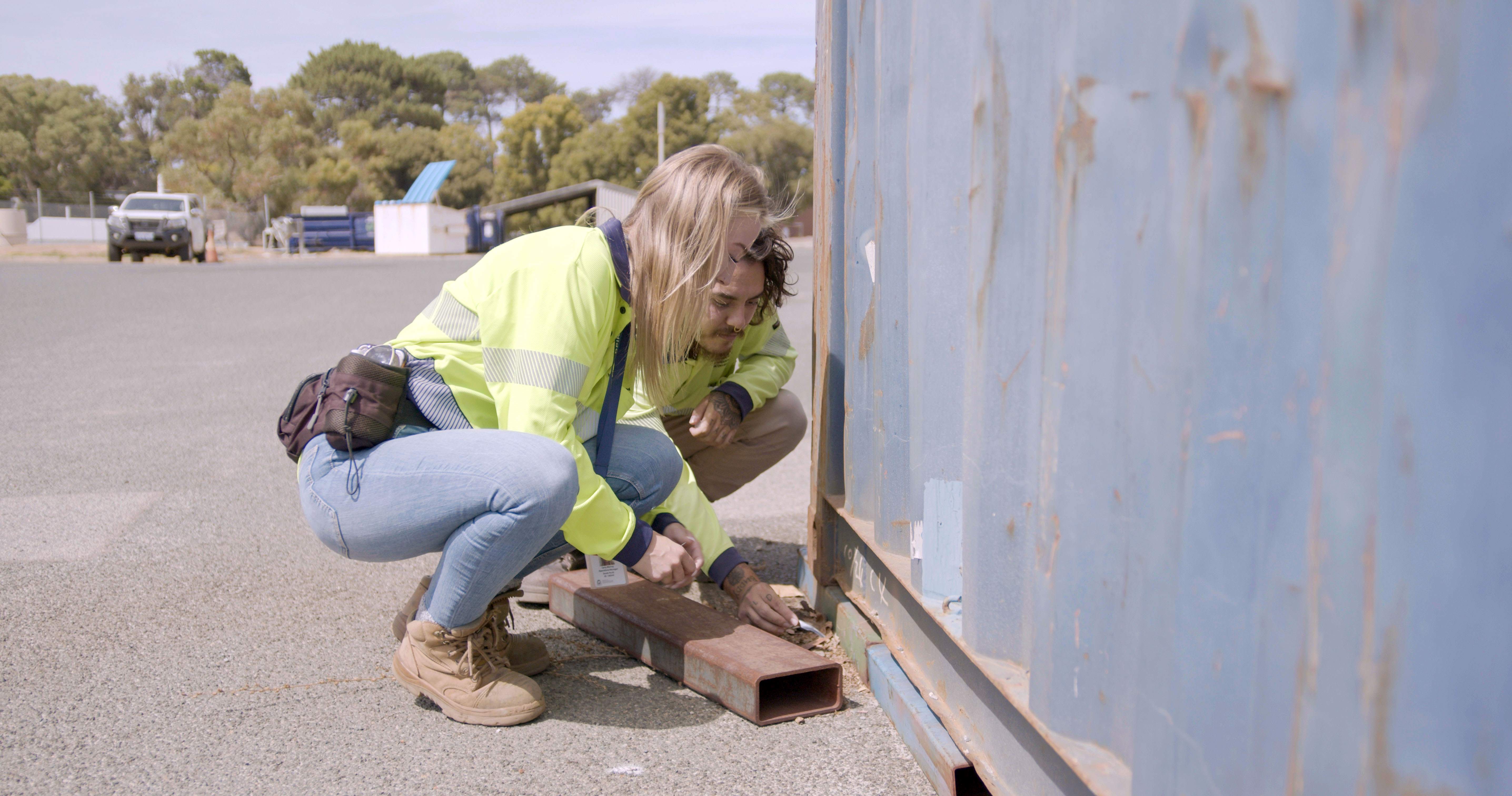 A woman and a man crouched near a cargo container.