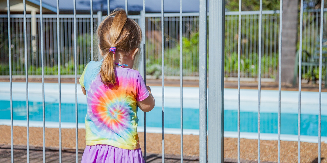 This image depicts a young girl standing at a swimming pool gate. 