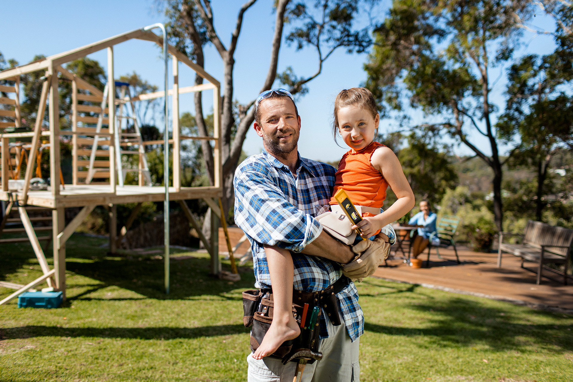 A man in a plaid shirt and tool belt holds a young girl in an orange top, with a construction project in progress behind them in a sunny outdoor setting.