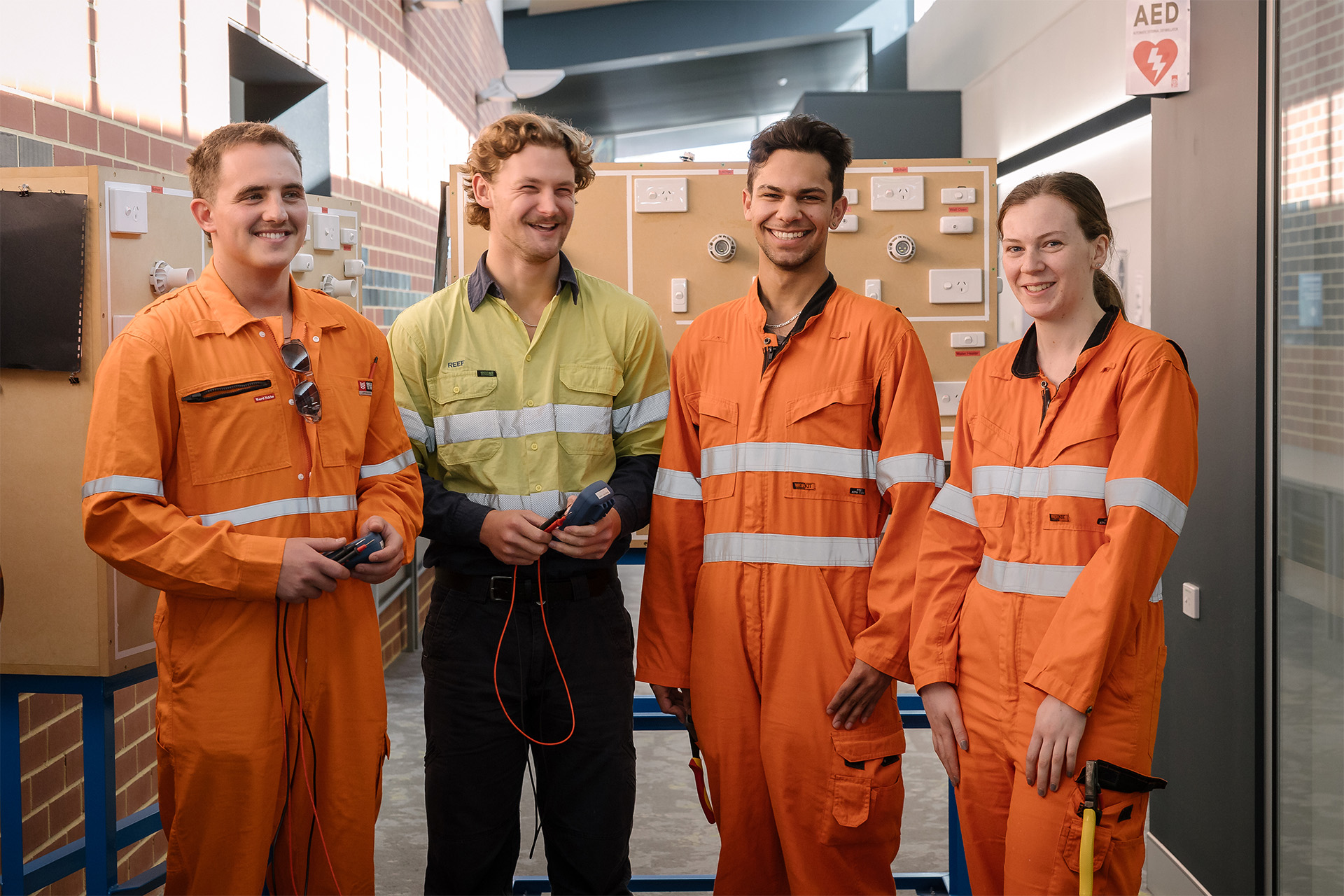 Four students in safety uniforms smiling in a training facility with electrical equipment in the background.