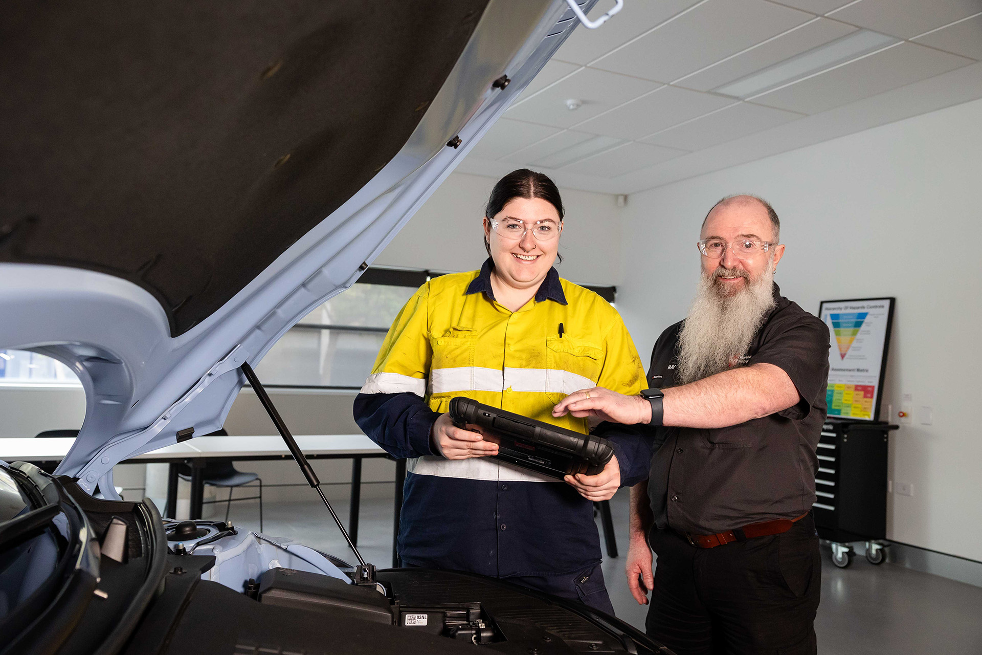 A woman in a high-visibility work shirt holds a diagnostic tool while standing beside an older man with a beard. They are inspecting an open car hood together in a workshop setting.