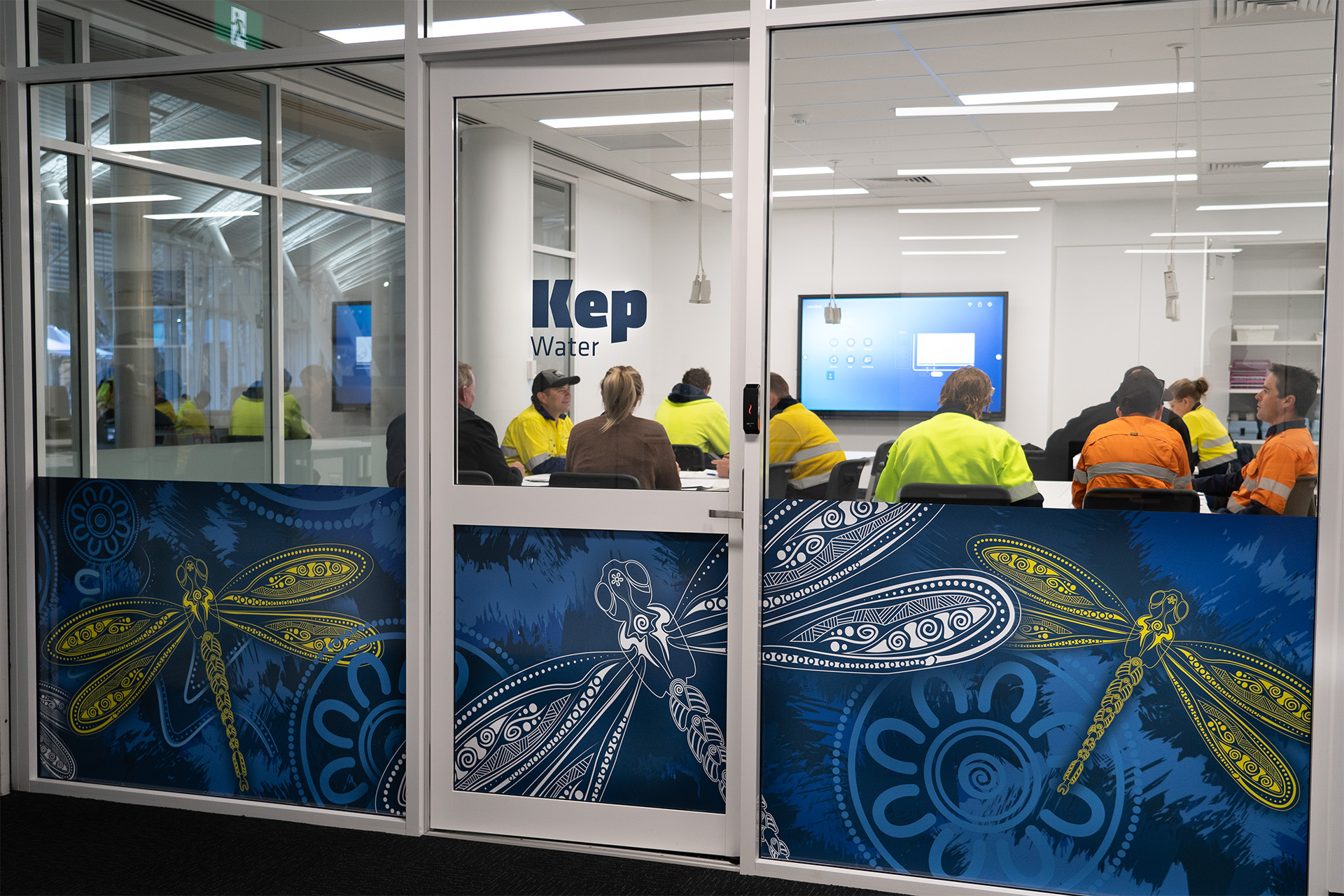 People in high-visibility workwear attend a meeting inside a glass-walled room labeled 'Kep Water,' with large decorative dragonfly artwork on the lower panels of the glass.