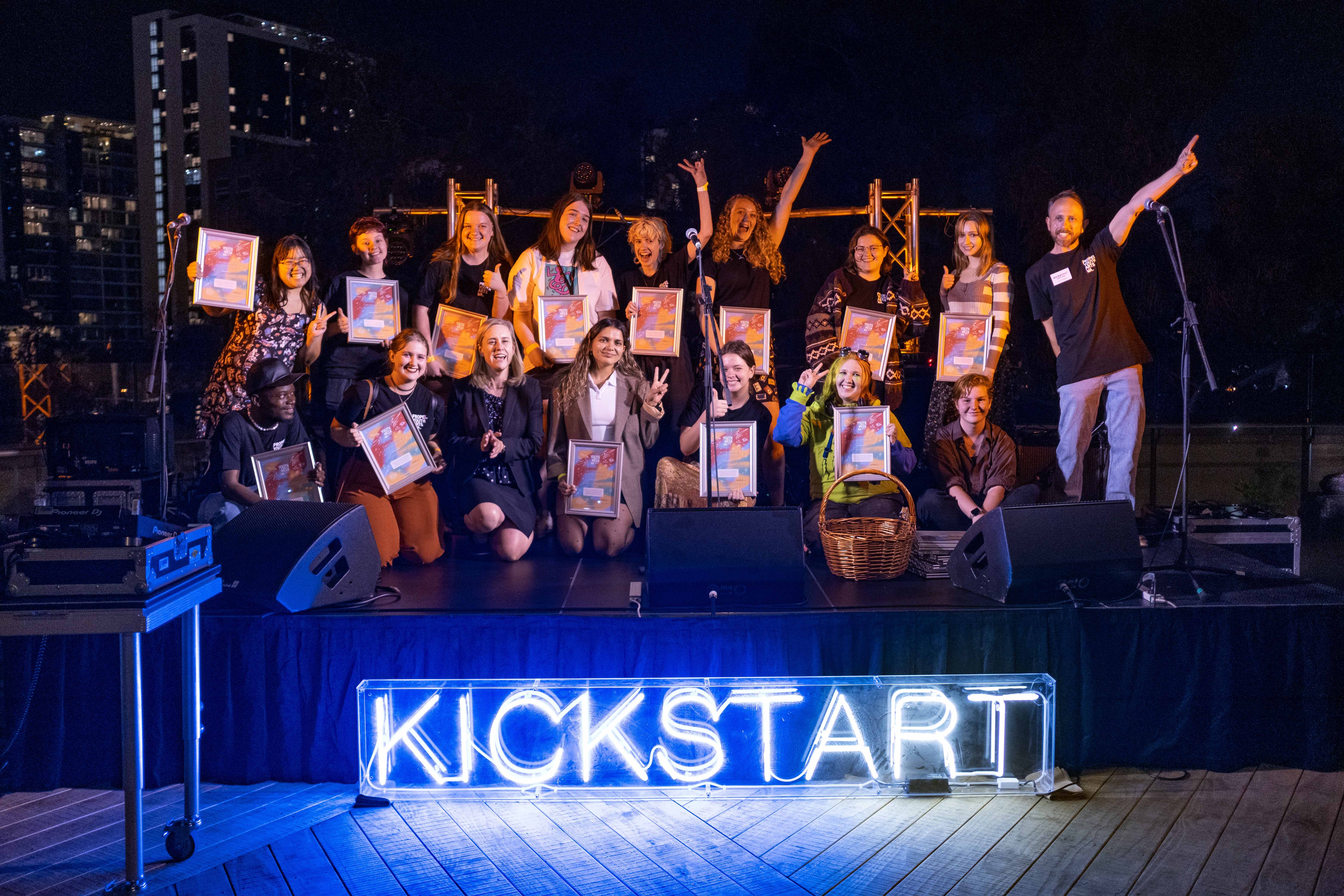 Group photo of 16 young adults on stage with the word Kickstart lit up in front of the stage