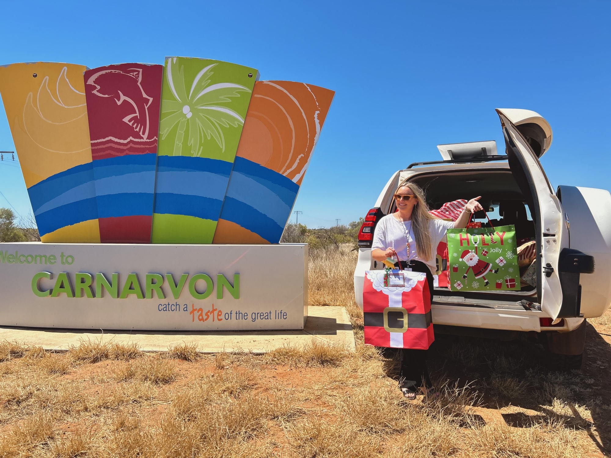 Image of a woman with a car full of Christmas presents next to the welcome to Carnarvon sign