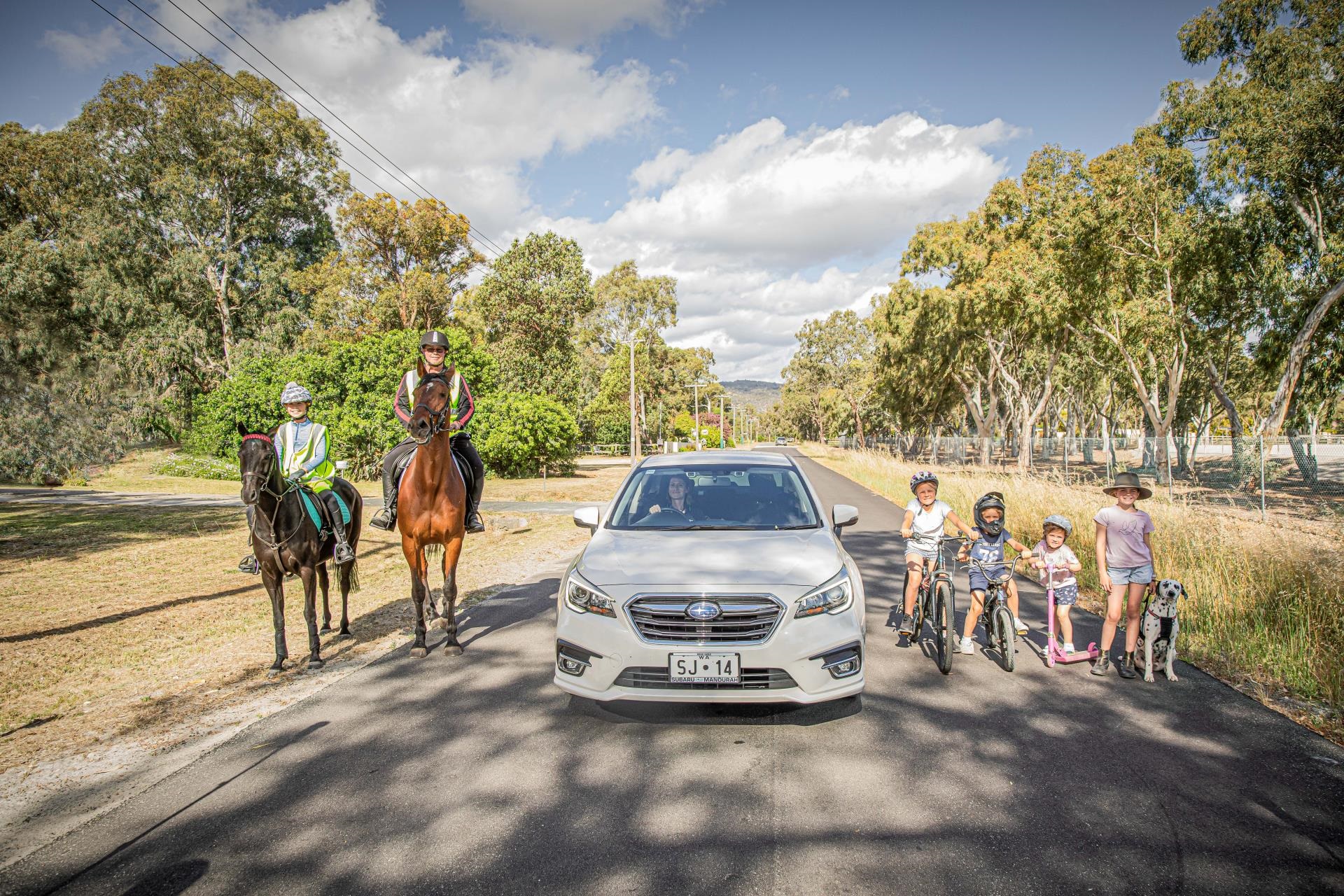 A car on the road flanked by two horses on the left and four kids on the right