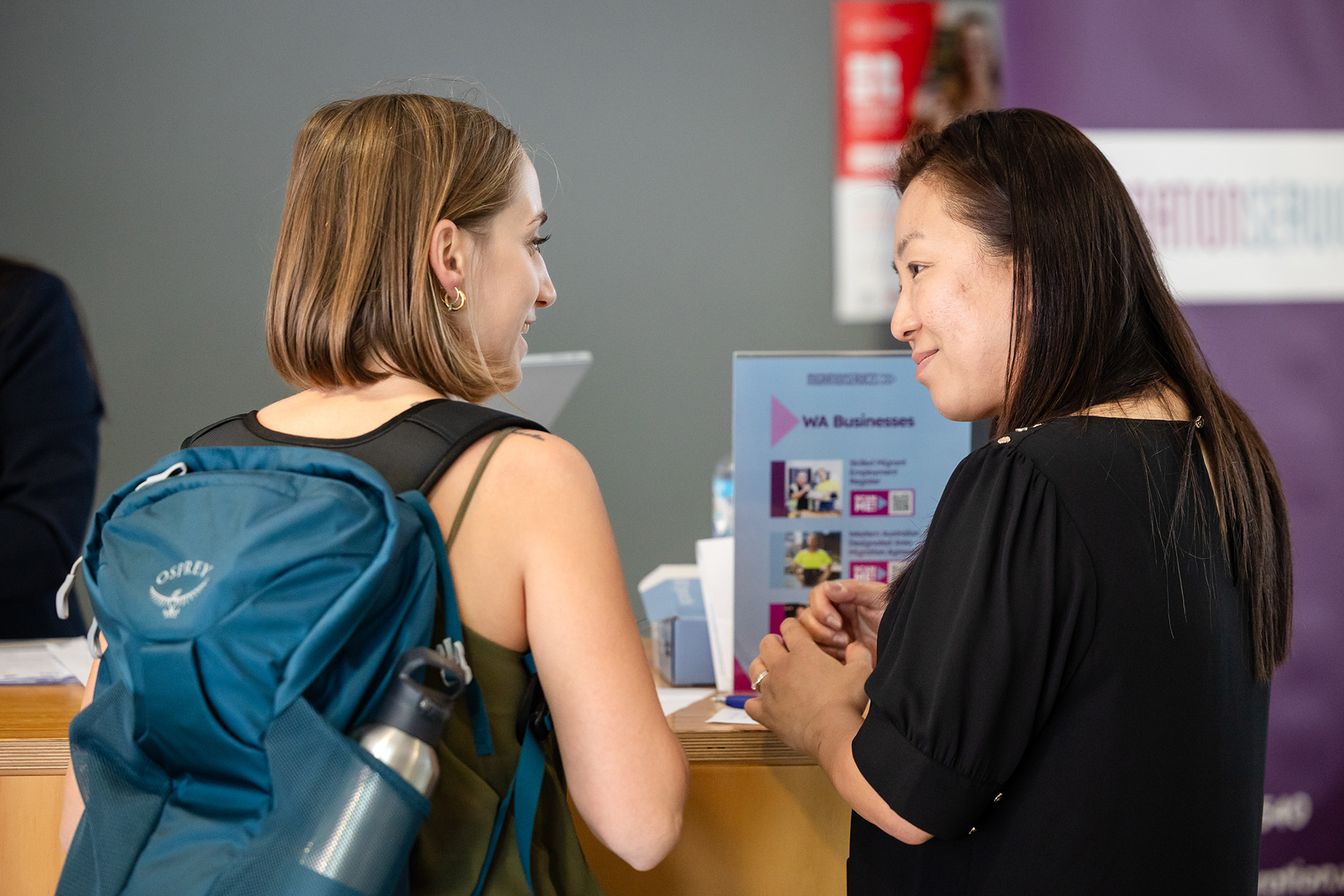 Two women interacting at an information desk, one with a backpack and the other providing details with a smile.