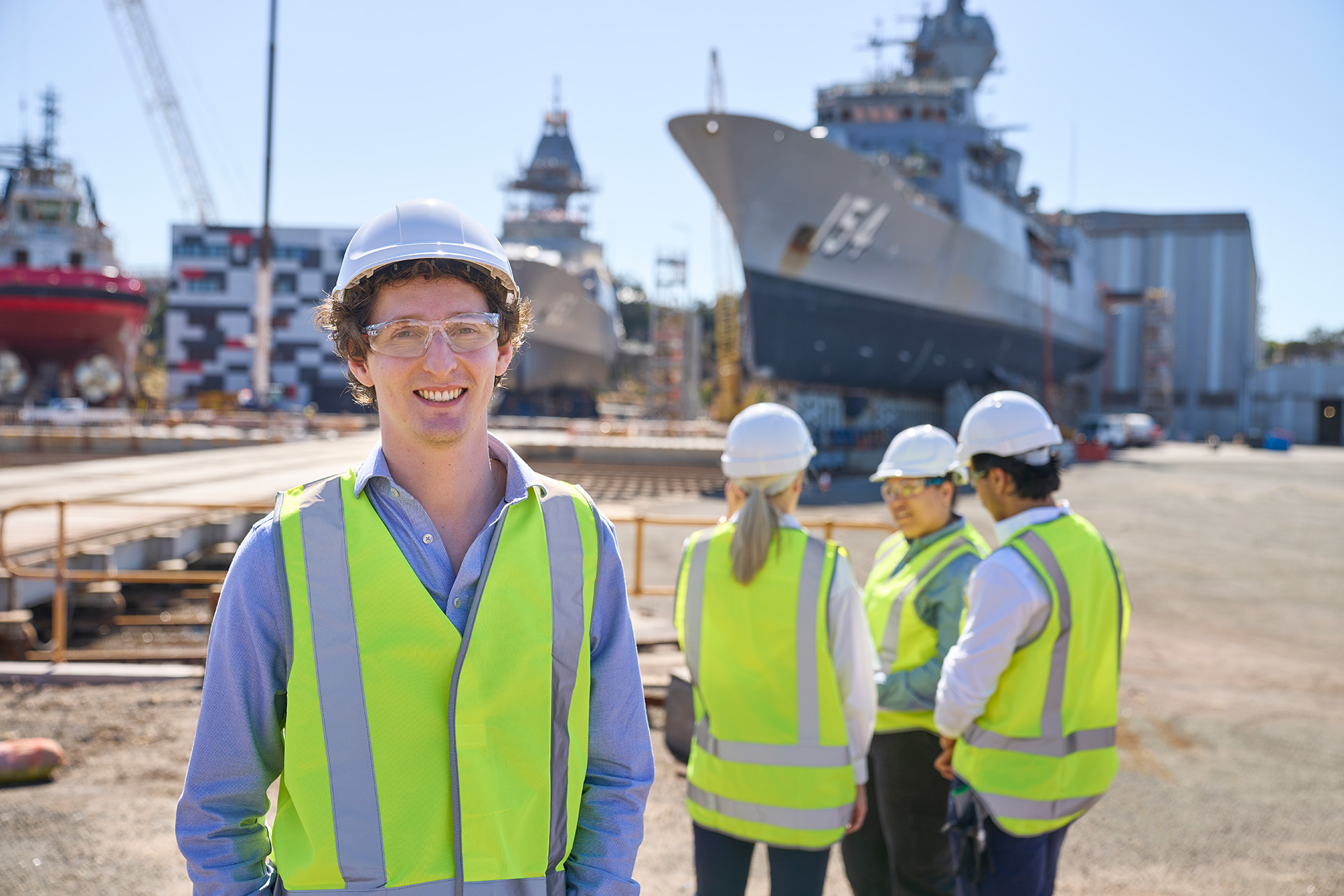 Person in a high-visibility vest and hard hat smiling in a shipyard, with colleagues and large ships in the background.