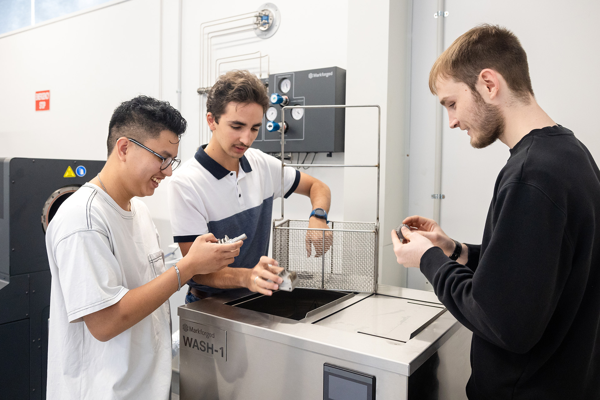 Three young individuals examining 3D-printed components near a Markforged Wash-1 machine in a technical lab setting.