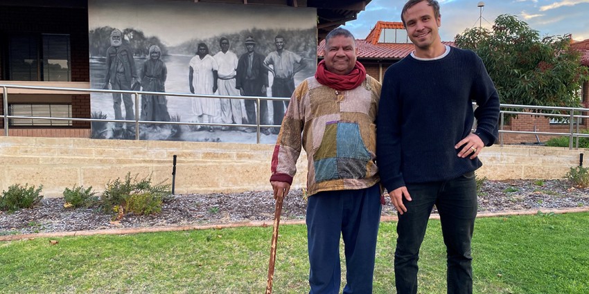 Aboriginal custodian Joe Northover stands on the grass at the front of the Collie Shire Offices with a cane in hand alongside mural artist Jacob 'Shakey' Butler, with the new Throssell St black and white Aboriginal Mural behind them