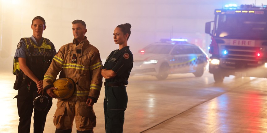 a police officer, a DFES officer, and a paramedic stand in front of some emergency vehicles. The vehicles are a fire truck with its lights on and a police car