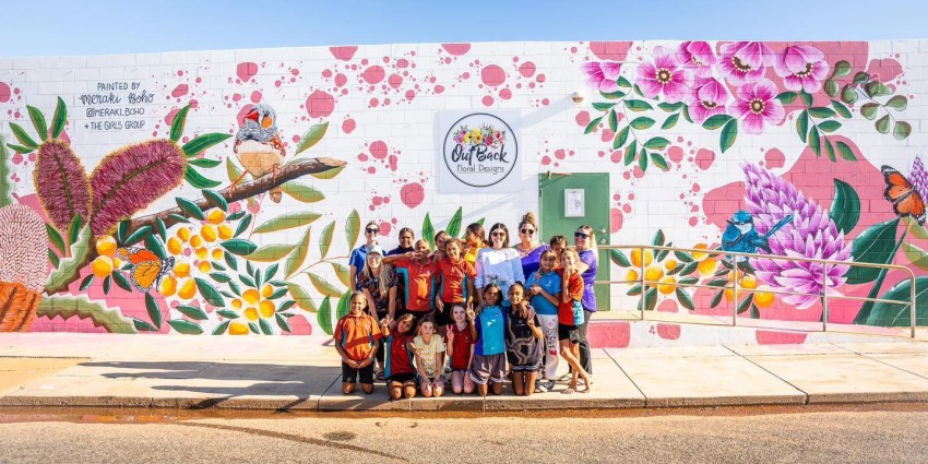 group photo of a group of Aboriginal girls and women on a street with a colourful murel on a wall behind them