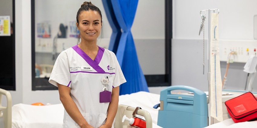 A female nursing student smiling in a hospital room, wearing a white and purple uniform.